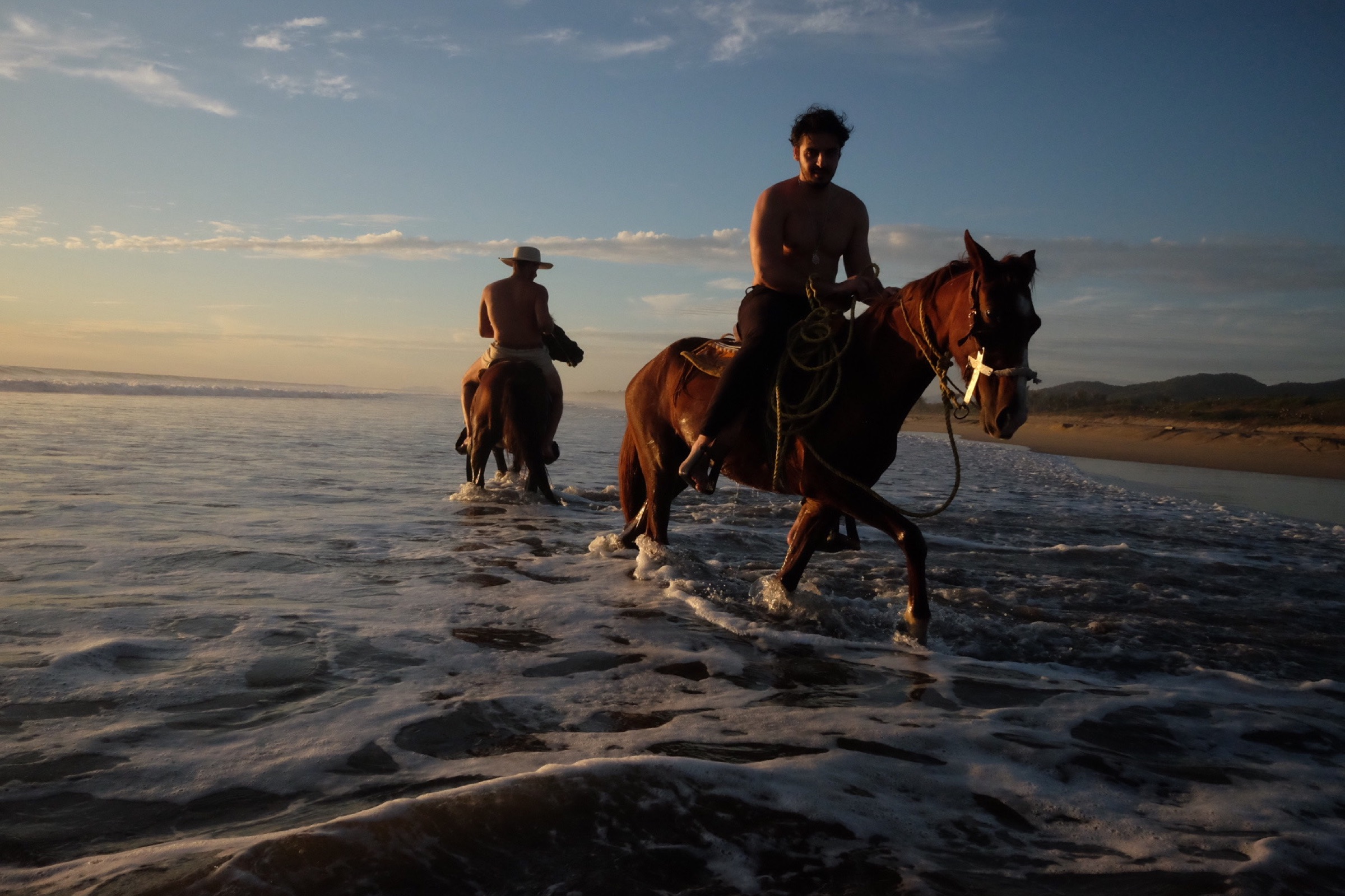 Two riders on horseback wading through the surf toward the shore in morning light